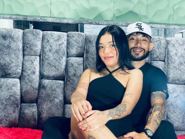 SachaAndFercho sitting on a gray, textured couch. SachaAndFercho has black hair, tattoos, and wears a black, halter dress. Fercho has a beard, tattoos, and a white cap.
