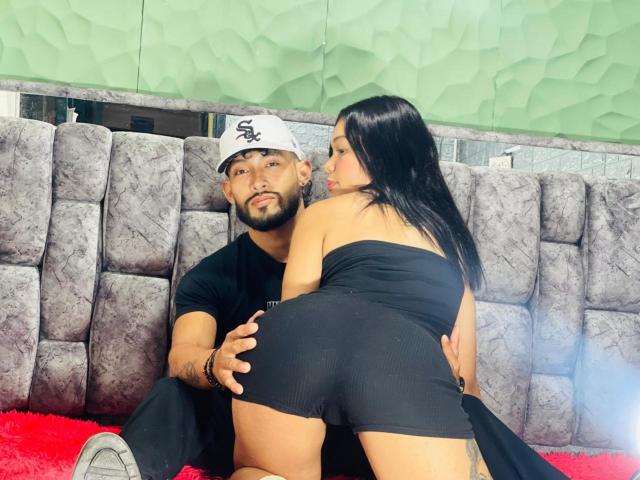 SachaAndFercho, a bearded man in a black shirt and white cap, holds Fercho's black ass, who's in a black strapless top, kneeling on a red rug.