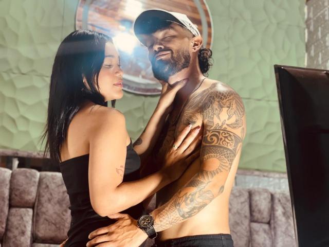 SachaAndFercho, shirtless with tattoos, wears a cap, standing, arms around Fercho's chest, who's in a black dress, hands on Sacha's chest, both smiling, light flares in front.