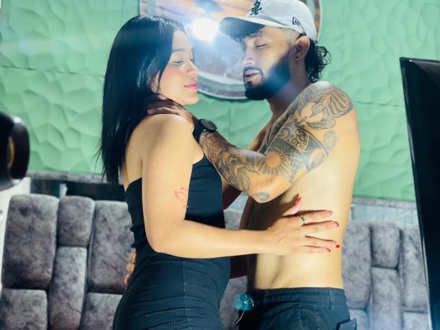 SachaAndFercho with black hair, wearing a black dress, holding and kissing a tattooed, shirtless Fercho with a cap, and black pants, in an intimate, warmly lit moment.