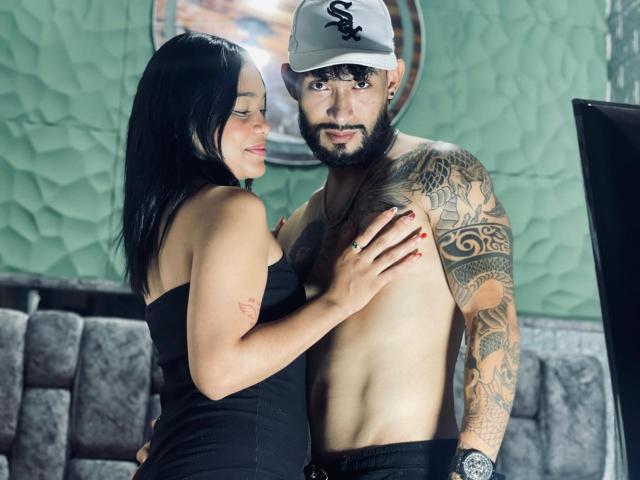 SachaAndFercho, with dark hair and red nails, touches Fercho's tattooed chest and abs. Fercho, bearded, wears a cap, exposing his shirtless torso. She's in a black strap dress.