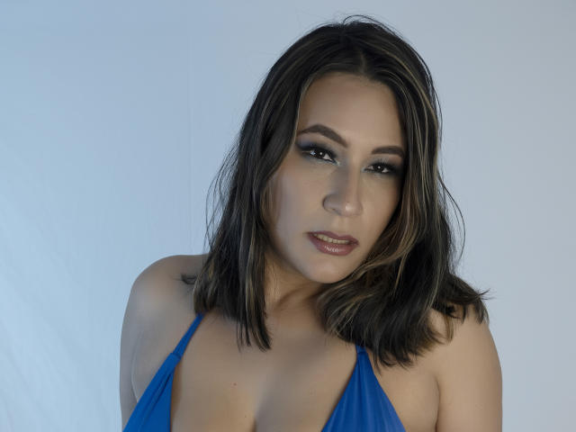 EmaFoxy with medium brown hair, blue halter top, showing her tits, slightly parted lips, looking down, subtle makeup, soft light, white background, medium skin tone, attractive.
