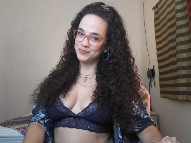 AmyFlores, met krullend donker haar, draagt een bril, een blauwe kanten bh, een open shirt en een ketting. Ze glimlacht naar de camera en laat haar borsten zien, met blauwe oorbellen en een tatoeage op haar arm..