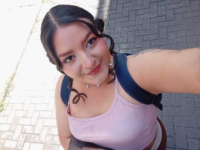 VictoriaRiver with curly black hair, pink tank top, and black backpack, smiling at the camera, showing her tits and tattooed arm, standing on a brick sidewalk.