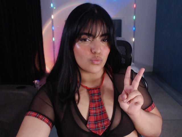 Zafirojh with black hair, wearing a sheer black top with a red plaid necktie, making a peace sign, pouting with a slight smile, in a blue-walled room.