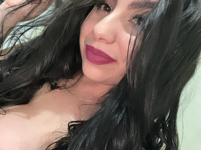VenusxMusa with long black hair, purple lipstick, and brown eyes, smiling at the camera, showing her bare shoulders and a hint of her tits.
