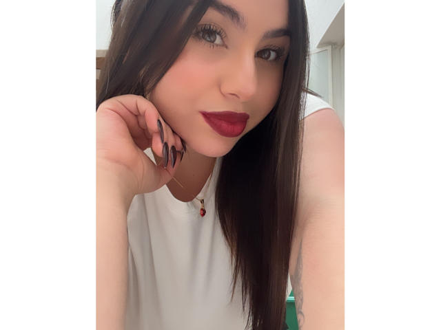 VenusxMusa has long brown hair, red lipstick, black nails, white shirt, and a necklace with a red gem. She's leaning on a hand, looking at the camera.