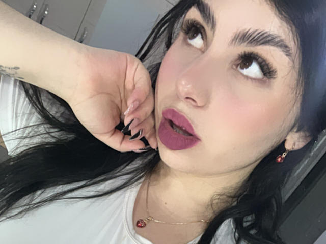 VenusxMusa with long black hair, dark eyebrows, and pink lipstick, looking sideways with her hand near her mouth, wearing a white shirt and red earrings.
