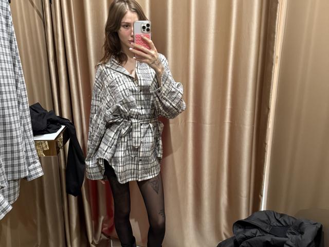 SensualMila with wavy brown hair, wearing a gray plaid dress, black tights, and holding a pink phone, showing her tattooed thigh in a dress store.