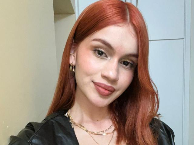 MarilynScotty with red hair, wearing a black leather jacket, gold necklace, and hoop earrings, smiling with pink lipstick, and a subtle tan. Boobs are not visible.