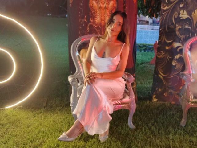 MindyLove sits on an ornate, vintage chair with a floral pattern. She wears a flowing, white, spaghetti-strap dress and white heels. Her long brown hair is loose, and she looks relaxed. A circular light source is on the left, casting a warm glow. The background features dark, patterned panels and a grassy area. MindyLove's dress has a subtle sheen, and her pose is casual. The overall composition is balanced, with the light source and patterned panels creating visual interest.