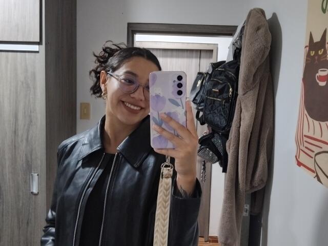 Mllevv with dark hair in a messy bun, glasses, and a black leather jacket, smiling while taking a selfie with a purple phone, showing a cute face and ass.