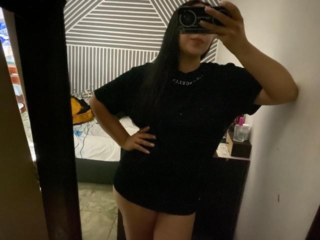 CherryDamore with long black hair, black t-shirt, and no pants, standing in a room, taking a mirror selfie with one hand on her hip, showing her bare ass.