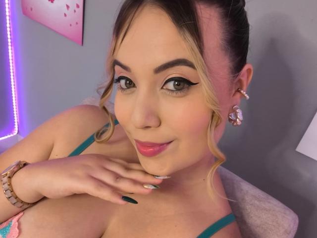 ZoeTobon69 has pink lipstick, green eyeliner, and pink earrings, with blonde hair in a bun, green nails, and a pink bracelet, showing her large tits and smiling.