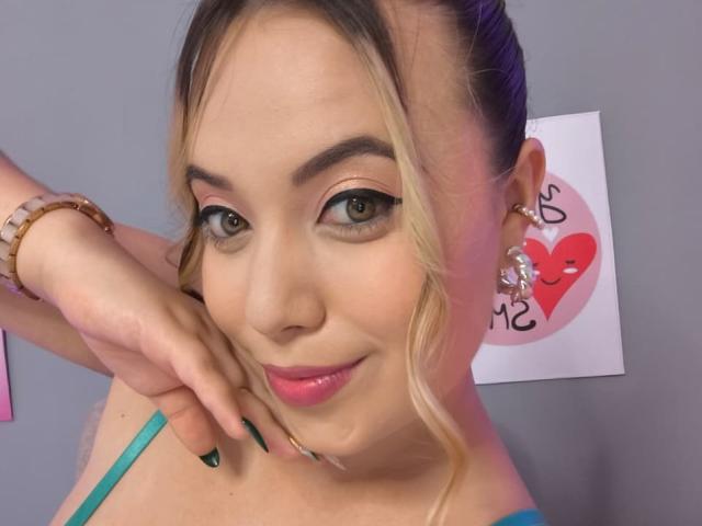 ZoeTobon69 with blonde hair in a bun, green eyes, and pink lipstick, wearing a blue top, and a wooden bracelet, has a hand on her cheek, smiling.