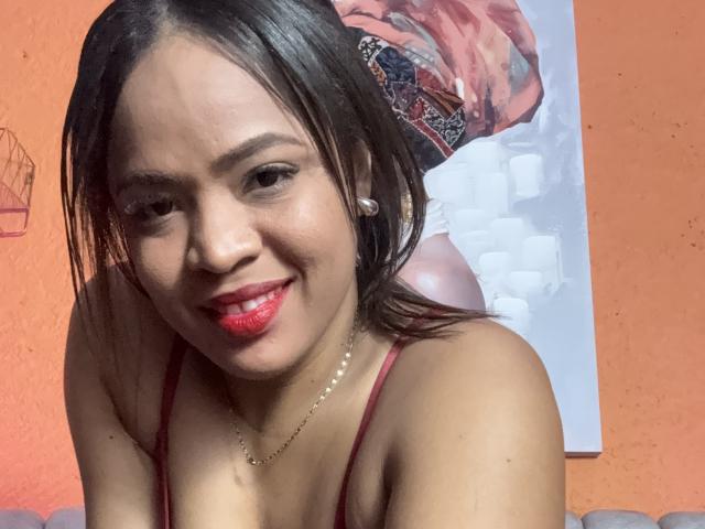 AliceBooty tiene el cabello castaño oscuro, lápiz labial rojo y un collar, sonriendo con tirantes rojos de espagueti y aretes, mostrando sus pequeños senos y un rostro lindo..
