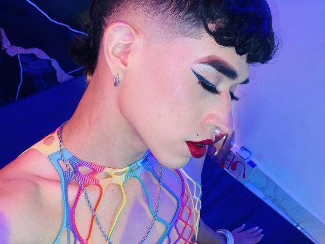 BriggitteLove with short curly black hair, pale skin, bold red lipstick, and colorful netted top, showing off her nose ring and hoop earrings, eyes closed, looking down.