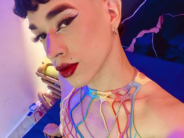 Close-up photo of BriggitteLove with short, curly dark hair and bold, winged eyeliner. She has dark red lipstick, a small beauty mark under her right eye, and a small black stud in her left ear. BriggitteLove wears a colorful, net-like top with rainbow patterns. Her right hand holds a yellow roll of paper. The lighting is purple and blue, highlighting her skin and makeup. The image has a vibrant, modern aesthetic. Slight shadow on her face.