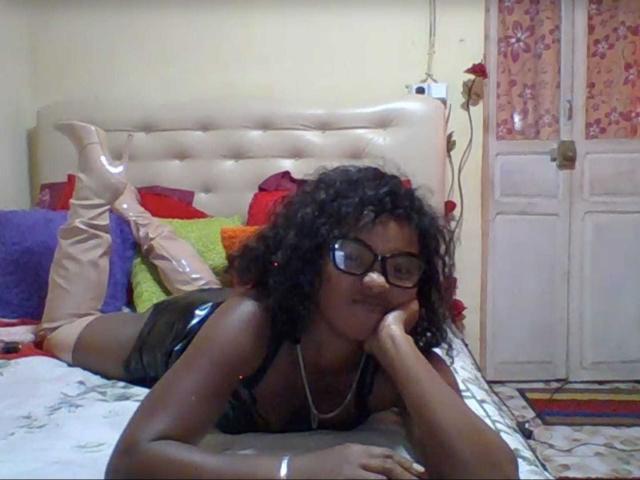 Maryssah, avec ses cheveux noirs bouclés, ses lunettes et sa peau foncée, est allongée sur un lit, vêtue d'un débardeur noir, de bottes beiges montantes et d'un collier, et repose son menton sur sa main..