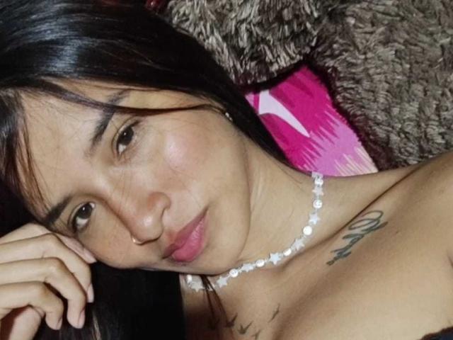 ImmyAllison with long black hair, wearing a beaded necklace, has pink lips, brown eyes, and a tattoo on her right chest. Her hand rests near her face.