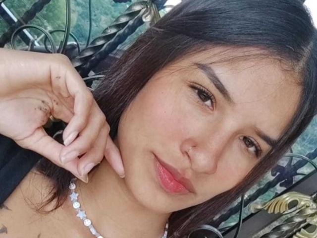ImmyAllison with dark hair, nose ring, subtle makeup, pink lips, wearing a pearl necklace, hand near face, slightly smiling, brown eyes, tattoo on finger, casual selfie.