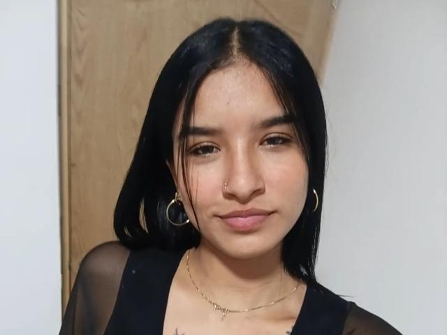 ImmyAllison with straight black hair, brown eyes, small nose ring, gold hoop earrings, black sheer top, subtle smile, light skin, and a delicate gold necklace.
