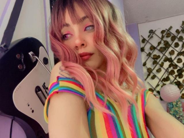 LaurenEvie69 with pink and blonde wavy hair, rainbow-striped shirt, pink eyeshadow, and nose ring, holding a black and white guitar. Her tits are small, she's cute.