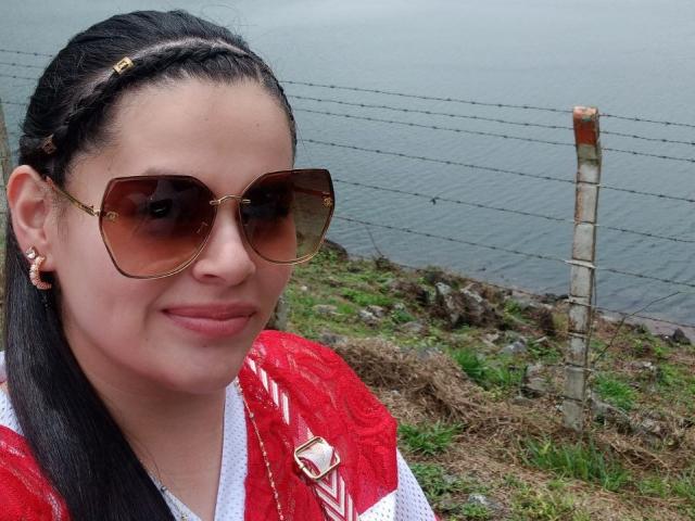 ValeriaNoir with braided black hair, brown aviator sunglasses, red and white sports jersey, and earrings, smiling near a wire fence and grassy area. Her tits are small.