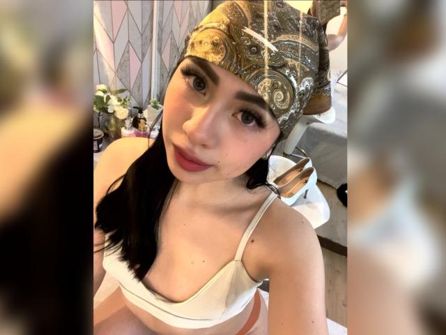 ZoeRedds with dark hair in a white bra, gold patterned headpiece, and red lipstick, showing off her small tits and cute face in a selfie.
