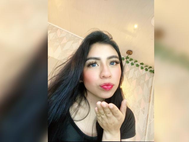 ZoeRedds with long black hair, blue eyes, pink lipstick, black shirt, and fair skin, pouting lips, hands near chin, looking at camera, soft light, slightly blurred foreground.