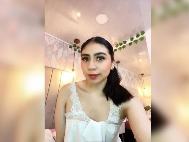 ZoeRedds with dark hair in a ponytail, wearing a white lace-trimmed dress, showing small tits, and a cute smile, in a softly lit room with hanging lights.