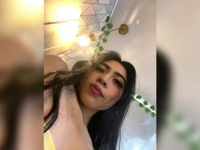 ZoeRedds, con cabello largo y negro, mirando hacia abajo a la cámara, sonriendo con lápiz labial rosa, mostrando parte de su escote con una blusa amarilla, bajo una lámpara de techo geométrica con plantas verdes..