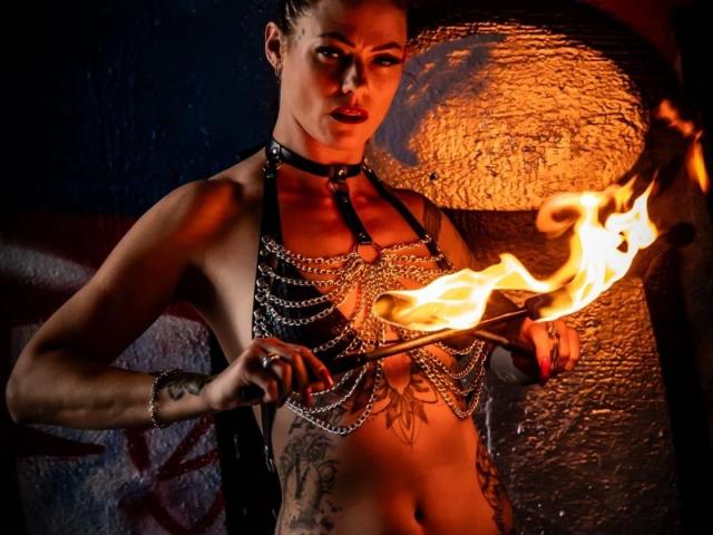 Sydney-Schmid-hot with tattoos, holding a flaming torch, wearing a black leather chest harness with chains, looking intense, in a dark, fiery-lit setting.