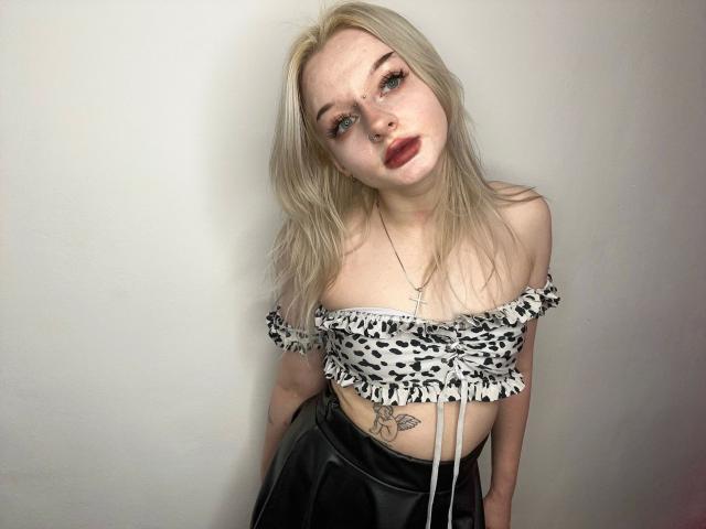 AlessaKiss-hot has pale skin, blonde hair, and blue eyes. She wears a black and white off-shoulder crop top with ruffled edges and a black, shiny, vinyl skirt. She has a tattoo on her right side below her ribs. Her lips are painted dark red, and she wears a cross necklace. She leans forward slightly, looking at the camera with a slightly tilted head. The image is clear and well-lit, with a simple, white background. Her expression is neutral, and her makeup is subtle.