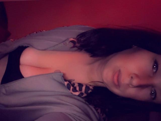 Siindyxx-hot with pale skin, dark hair, and blue eyes, wearing a leopard-print bra, grey shirt, and black panties, showing her boobs, lying down in red lighting.