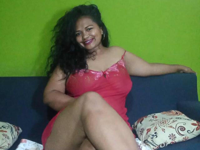 ChellaLove, a curvy woman with large tits, wearing a red lace nightie, sits on a blue sofa, smiling, legs crossed, with patterned pillows. Her dark curly hair is wavy.