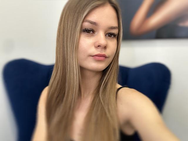 MidnightAura has long, straight, blonde hair, light skin, and a neutral expression. She's wearing a black top, with blue, heart-shaped chair in the background.