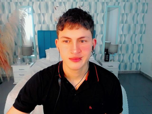 AxelSavage, ein etwas korpulenter Mann mit kurzen, braunen Haaren, der ein schwarzes Poloshirt und Kopfhörer trägt, sitzt in einem blau gestreiften Schlafzimmer und blickt mit einem leichten Lächeln in die Kamera..