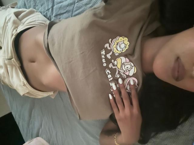 Babilett lying on a bed, wearing a brown shirt that exposes her midriff and beige shorts, with her hand on her chest, showing white-painted nails. Boobs visible.