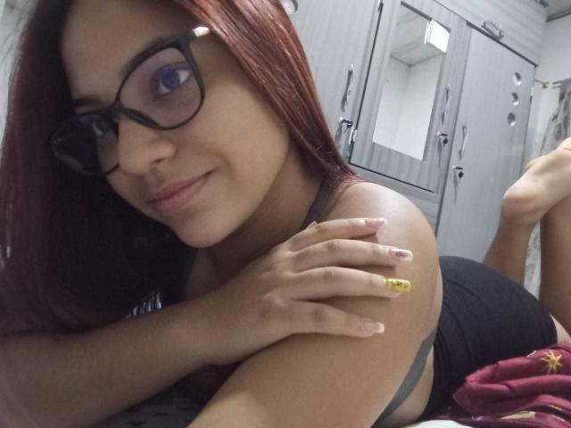 VeronicaBellerose with red hair, black glasses, smiling, lying on bed, black bra, yellow ring, hand on shoulder, legs up, bed with red and yellow blanket, bedroom.