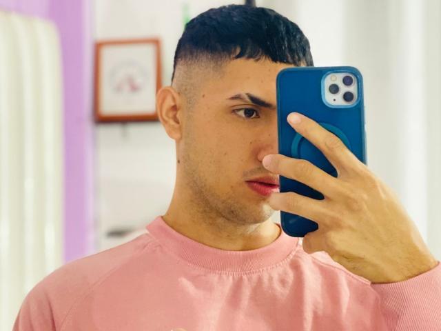 BaironSinclair with short black hair, wearing a pink sweater, taking a selfie with a blue phone, showing a serious face, and a slight beard. His nose is slightly red.