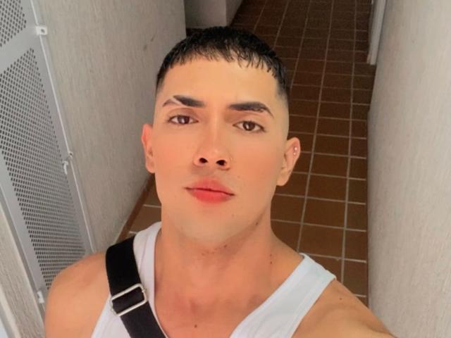 BaironSinclair has a serious expression, short dark hair, and is wearing a white tank top with a black strap. His brown eyes and pink lips are prominent.