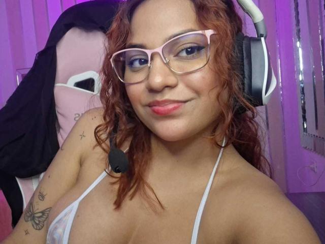 VeronicaBellerose with curly red hair, pink glasses, white headset, and a small tattoo of a butterfly on her arm, wearing a white halter top, showing her tits.