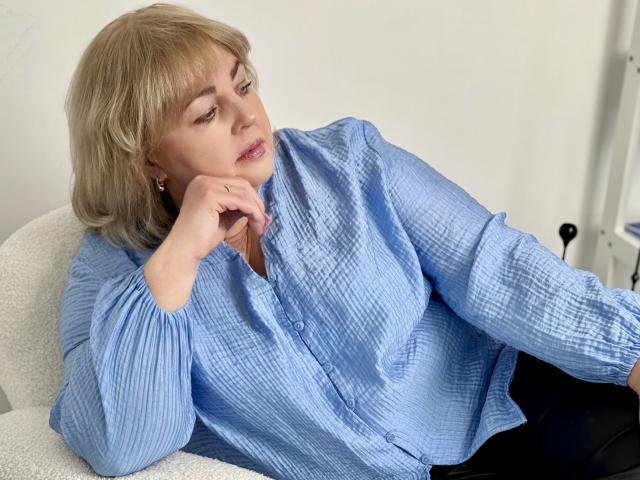 MilenaWendy with blonde hair, wearing a blue button-up shirt, black pants, and earrings, resting her chin on her hand, looking to the side. Her shirt is slightly wrinkled.
