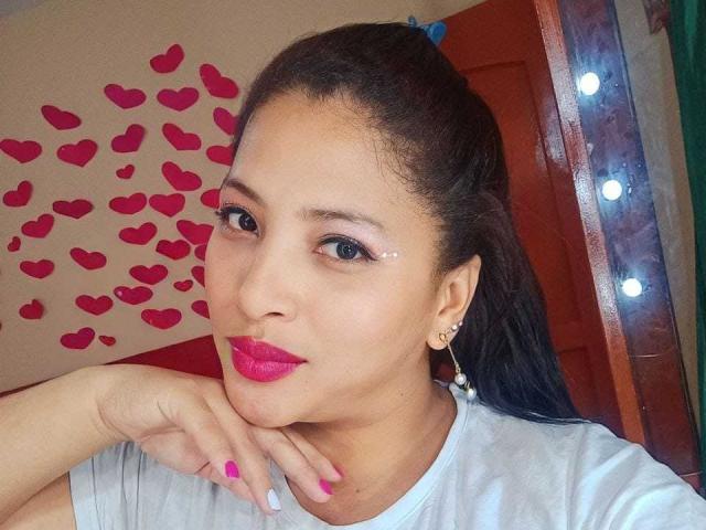 Roxane, een jonge vrouw met roze lippenstift, zwart haar in een staart, roze oogschaduw, een wit shirt, met haar hand op haar kin, roze nagels, parel oorbellen en een schattende glimlach, voor een spiegel..
