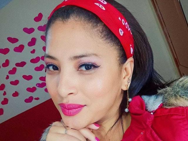 RoxaneMilf has pink lipstick, blue eyes, and a red bandana with white hearts. She's smirking, touching her chin, wearing a red jacket, and has pink nail polish.