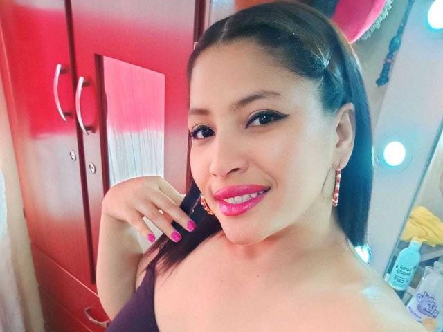 RoxaneMilf has pink lipstick, black eyeliner, and pink nail polish. She's smiling with her hand near her chin, wearing a black top, and has brown hair in a ponytail.