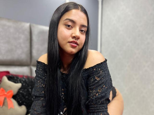 SalomeLee tiene el pelo largo y negro, una blusa negra de encaje con un corte que deja los hombros al descubierto, y un rostro con los labios fruncidos. Está sentada con las nalgas ligeramente levantadas, mostrando ropa interior negra. Su piel es de color marrón claro..