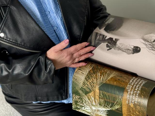 MilenaWendy's hand with pink nails touches a rolled-up magazine showing a black-and-white photo of a person on the sand. She's wearing a black leather jacket and blue shirt.