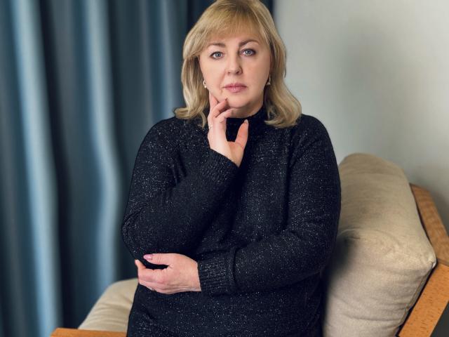 MilenaWendy with blonde hair, wearing a black sparkly sweater, sitting on a beige chair, hand on chin, looking serious, crossed arms, blue curtain in background.